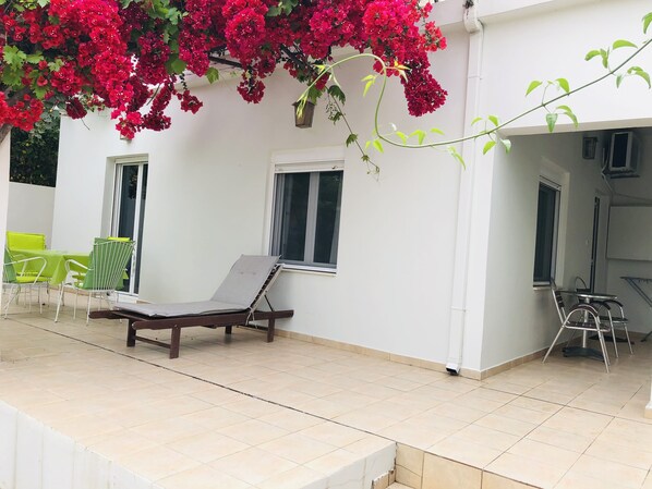 A covered outdoor area with a lounge chair, a table with chairs, and a view of a white building with windows.