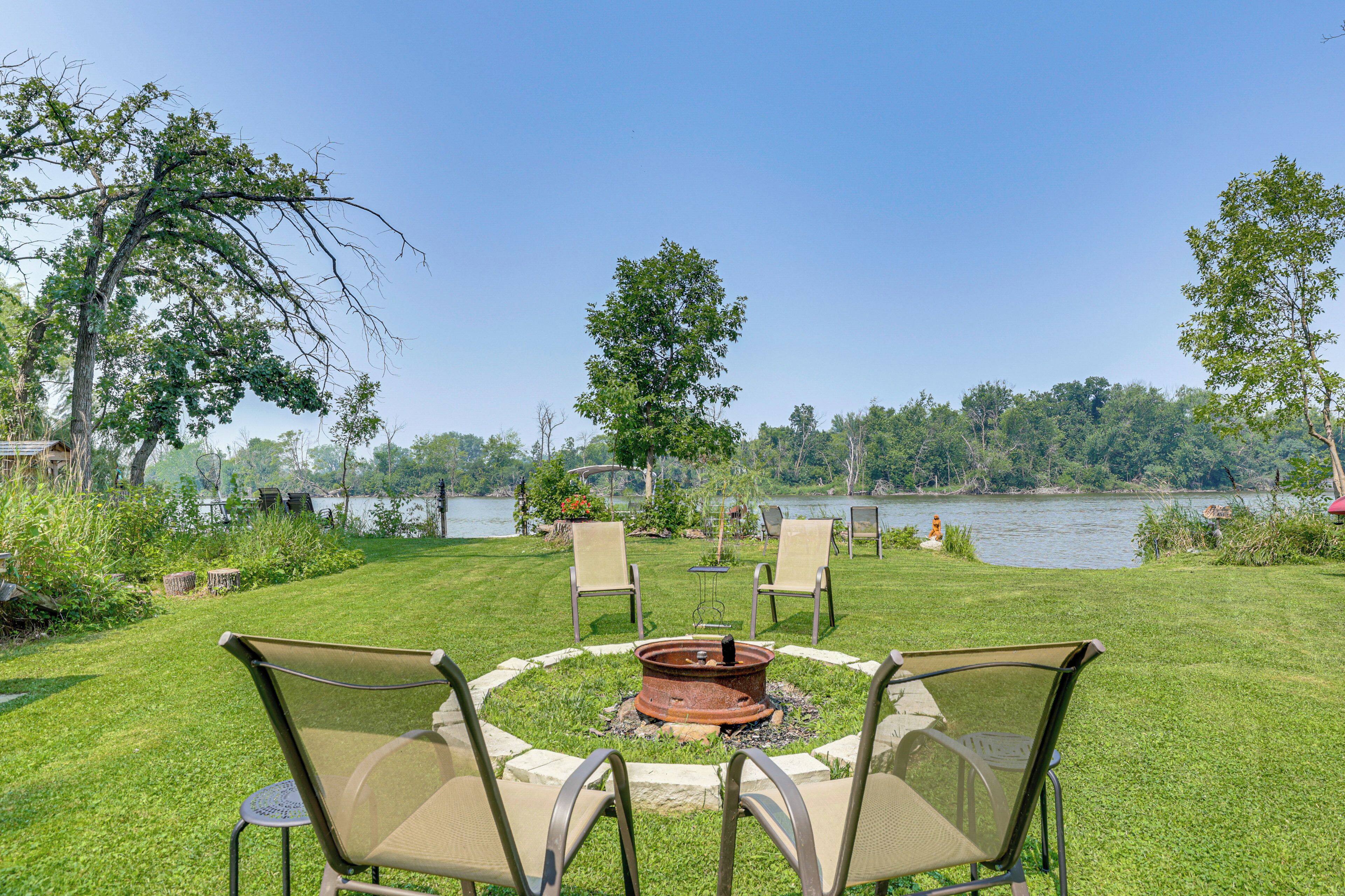 A circular outdoor seating area with a fire pit, surrounded by chairs, overlooking a lake.