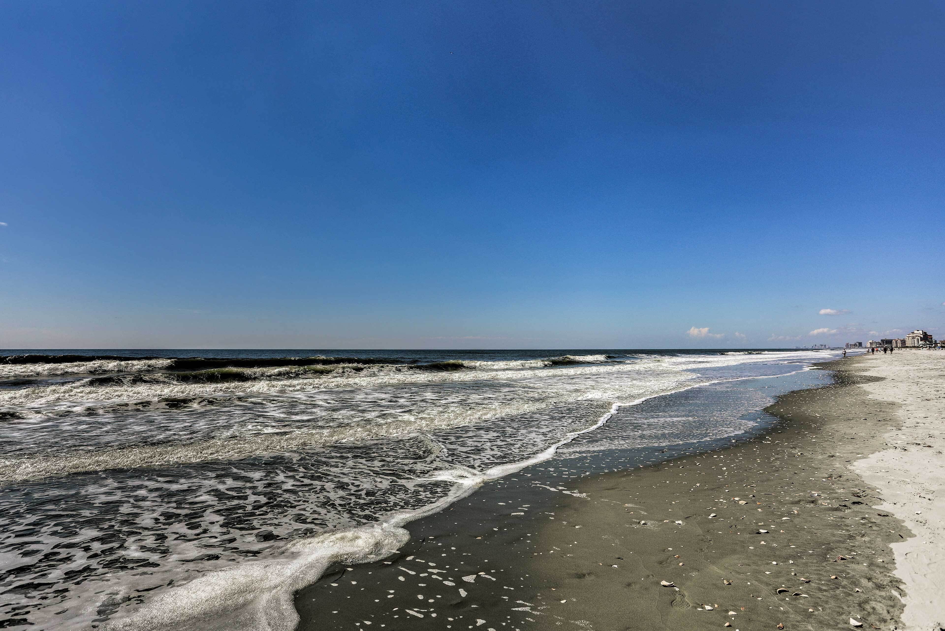 Une plage avec des vagues, un ciel dégagé et une côte lointaine parsemée de bâtiments.