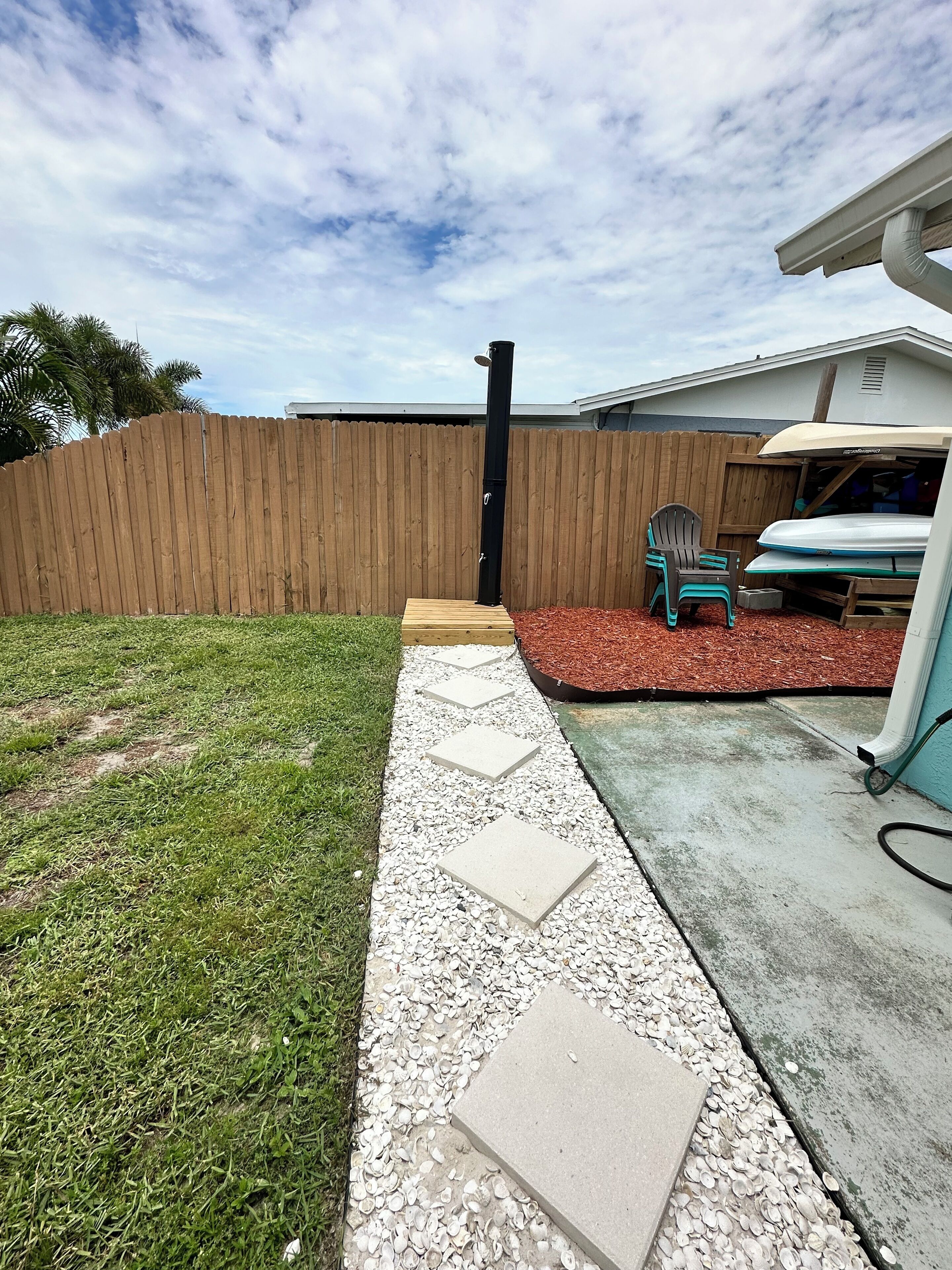 A backyard with a wooden fence, a concrete pathway, a patio area with a surfboard, and a small chair.