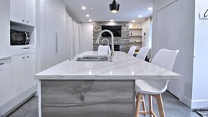 A modern kitchen with a large island, white cabinetry, a stone accent wall, and a flat-screen TV. - Peaceful Mountain Side Appartment Fully Equiped and Close to Many Activities (Bromont)