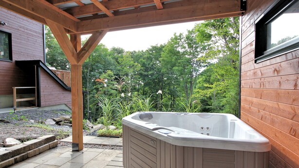 A hot tub in a covered outdoor area with wooden beams and a view of a forest. - Peaceful Mountain Side Appartment Fully Equiped and Close to Many Activities (Bromont)