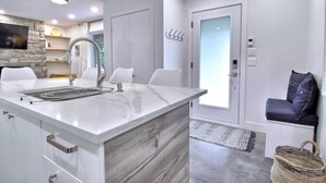 A modern kitchen with a marble countertop, a stainless steel sink, and a built-in oven. There is a white chair and a small table with a decorative item on it. - Peaceful Mountain Side Appartment Fully Equiped and Close to Many Activities (Bromont)