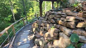 A stone pathway leading to a wooden deck with a railing, surrounded by greenery and trees. - Peaceful Mountain Side Appartment Fully Equiped and Close to Many Activities (Bromont)