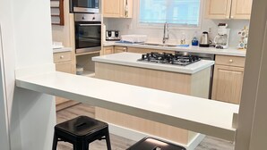 A modern kitchen with a central island, two black bar stools, wooden cabinets, a built-in oven, and a window with blinds. - Crystal Beach Getaway (Fort Erie)