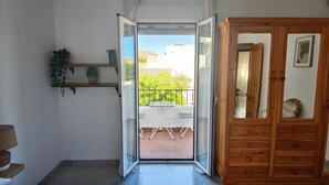 Een slaapkamer met een groot bed, een houten garderobe, een klein plankje met planten en uitzicht op een balkon met buitenmeubilair.