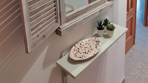 Een hal met een witte muur, een spiegel met houten lijst en een klein plankje met planten in potten.