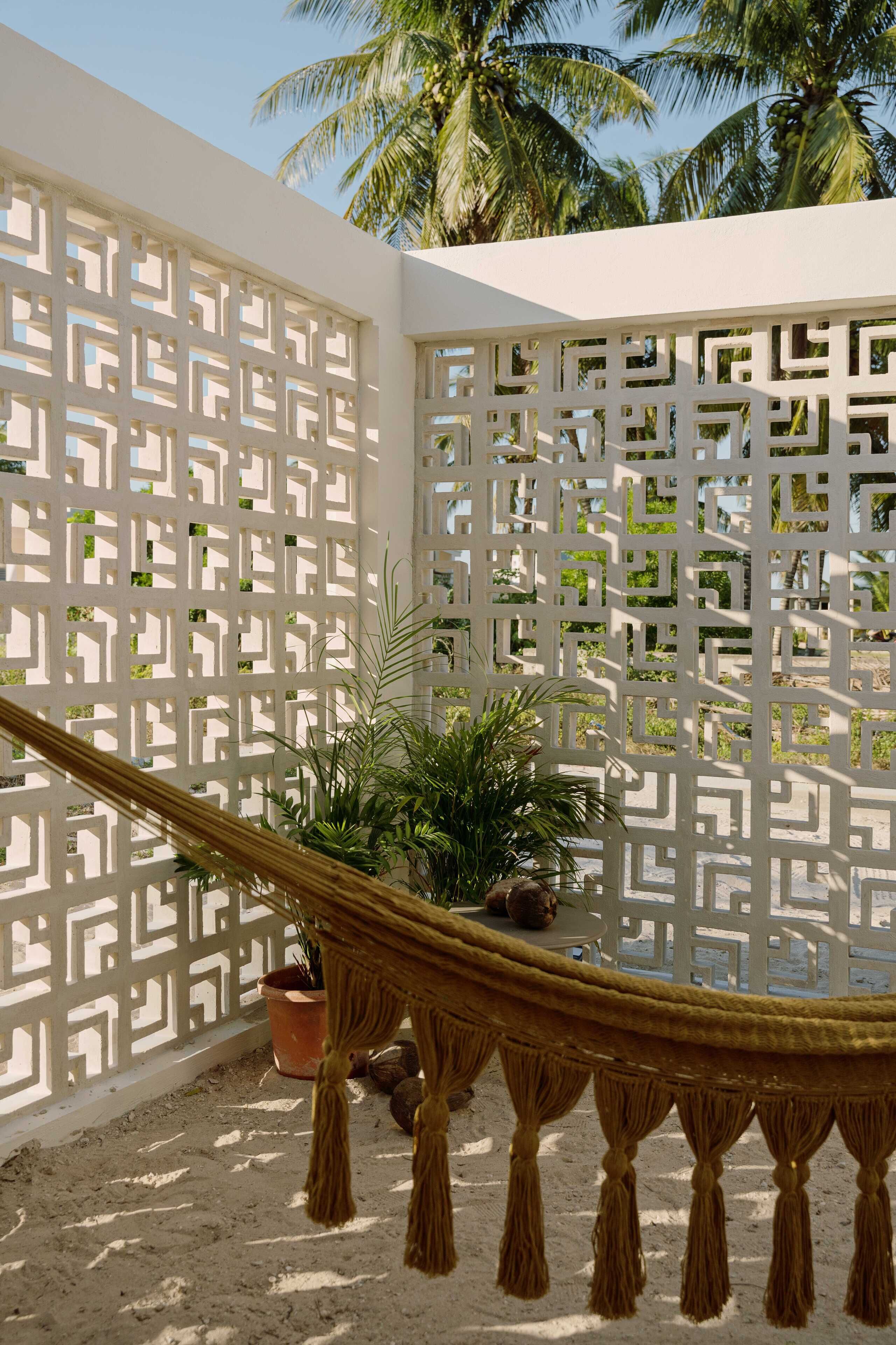 A hammock tied between white lattice panels, a potted plant, and a palm tree in the background.