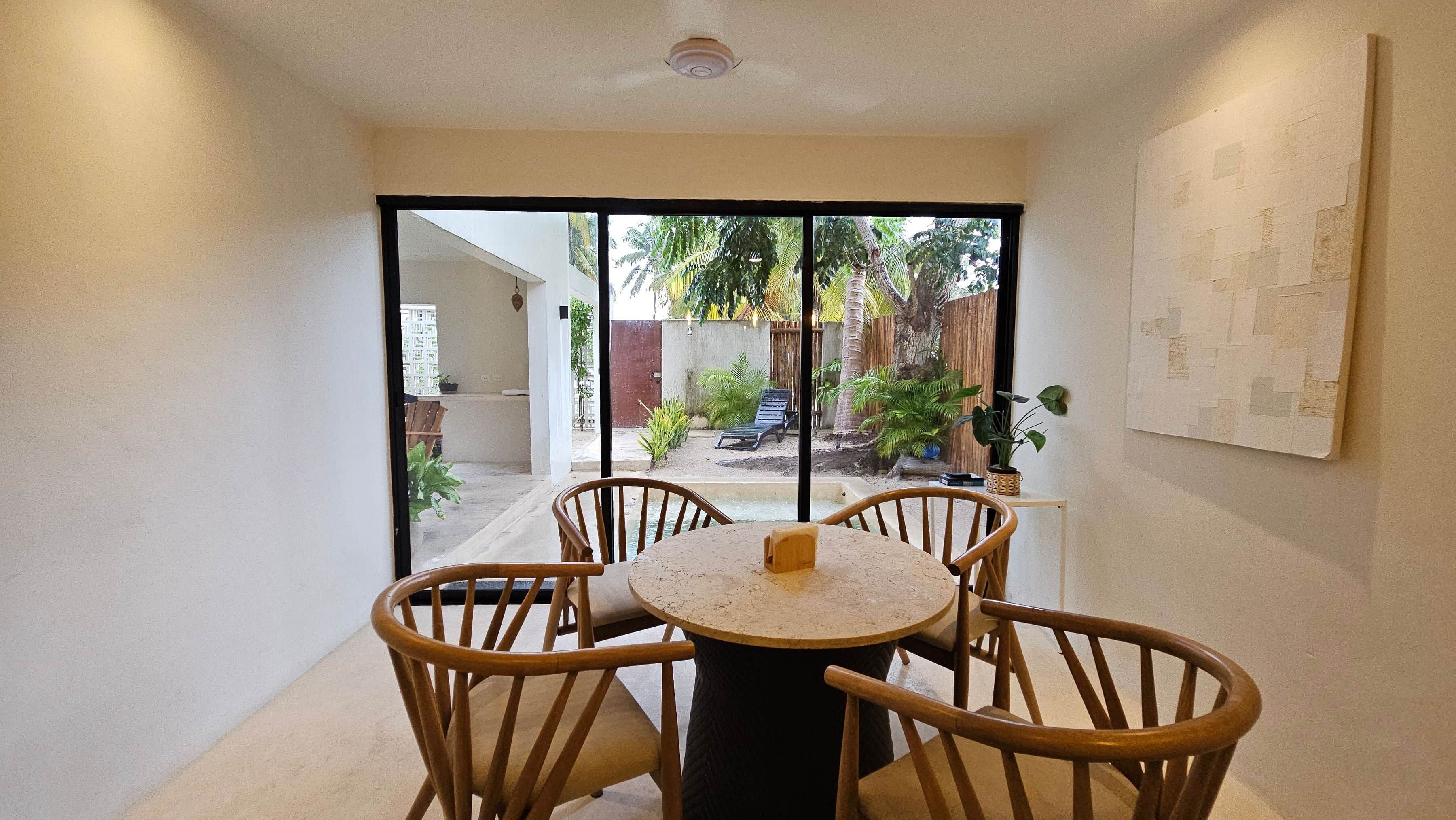 Una habitación con una mesa redonda y cuatro sillas de madera, vista a un jardín con plantas y una valla, y una pared con diseño decorativo.