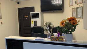 Un mostrador de recepción de hotel con una araña, un televisor en la pared, certificados enmarcados y una planta con girasoles.