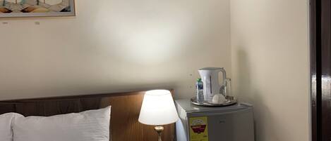 Una habitación de hotel con cama, mesita de noche con lámpara, refrigerador y un cuadro en la pared.