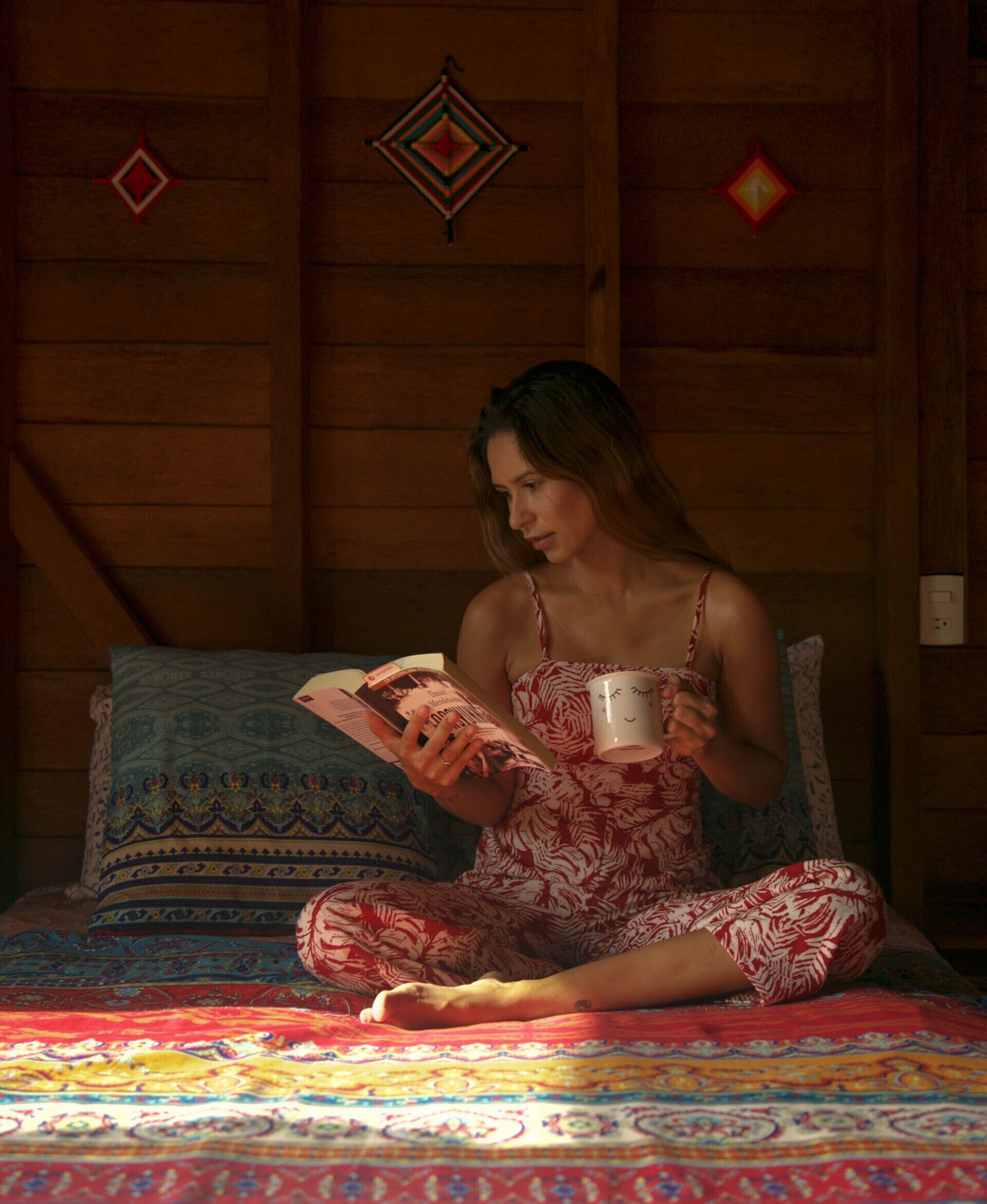 Una persona sentada en una cama en una habitación con paredes de madera, leyendo un libro y sosteniendo una taza.