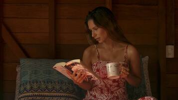 Una persona sentada en una cama en una habitación con paredes de madera, leyendo un libro y sosteniendo una taza.