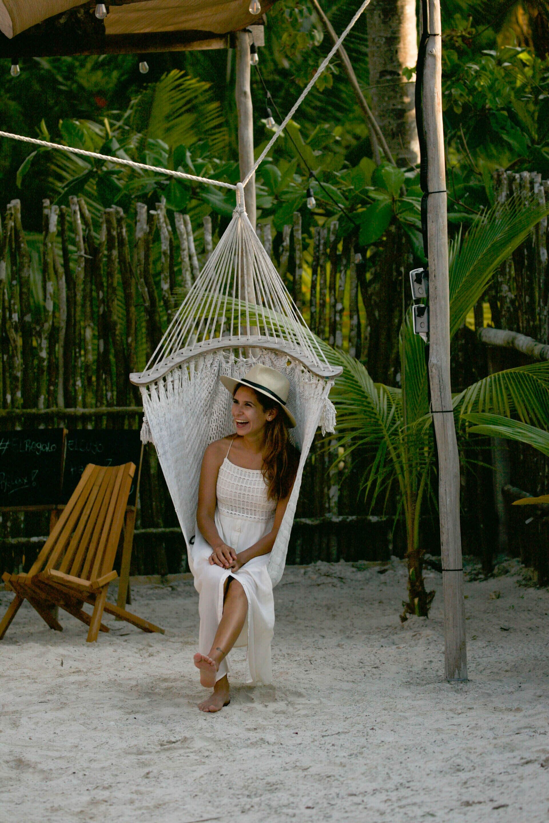 A hammock chair suspended by ropes, a wooden chair, and a signboard in a tropical setting.