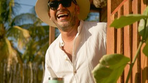 A person holding a drink, standing in front of a wooden structure with large green leaves. - El Regalo El Cuyo (El Cuyo)