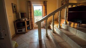Eine Treppe mit einem hölzernen Geländer führt zu einer Zwischenebene. Gegen die Wand steht ein Fernseher auf einem Holzgestell, und im Hintergrund ist ein Fenster mit Vorhängen zu sehen.