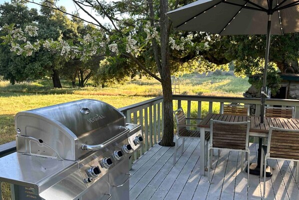 A barbecue grill on a wooden deck with a table and chairs, surrounded by trees and a grassy field.