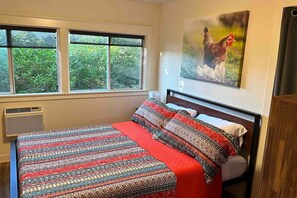 A bedroom with a large window, a bed with patterned bedding, a wall-mounted artwork of chickens, and a wooden headboard.