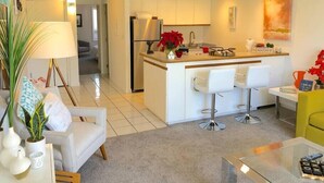 A modern kitchen with white cabinets, a central island with bar stools, and a dining area with a yellow chair. - Modern Beautiful 1BR Napili Bay Get-Away Condo (Lahaina)