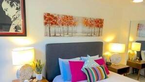 A hotel room with a large bed, multiple pillows, bedside tables with lamps, a painting on the wall, and a mirror. - Modern Beautiful 1BR Napili Bay Get-Away Condo (Lahaina)