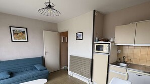 A compact living space with a kitchenette, a blue sofa, a white table, and a wooden dining table. - Pornichet - Studio Place du Marché (PORNICHET)