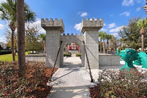 Enter through a grand stone archway, flanked by towers with crenellations, into a beautifully landscaped area with a patterned pathway.