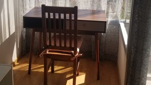 Un balcon avec une table et une chaise en bois, une fenêtre ornée de rideaux légers et une vue sur les immeubles.