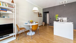 Ein moderner Wohnbereich mit Flachbildfernseher, einem Esstisch mit Stühlen, einem Bücherregal, einer Lampe und einer Küchenzeile inklusive Wasserkocher.