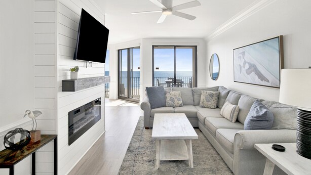 A modern living room with a flat-screen TV, a ceiling fan, a fireplace, a sofa, and a view of the ocean.