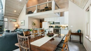 A spacious living area with a high ceiling, a dining table with wooden chairs, a kitchen with white cabinets, and a stone fireplace. - Ponderosa Pine Escape  (Truckee)