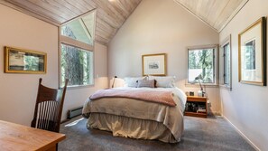 A bedroom with a large bed, a wooden chair, a desk, and multiple windows offering views of trees. - Ponderosa Pine Escape (Truckee)