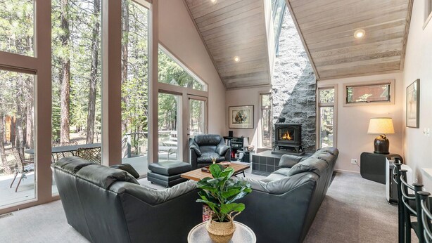 A spacious living room with a stone fireplace, leather sofas, and large windows offering a view of the surrounding forest. - Ponderosa Pine Escape  (Truckee)