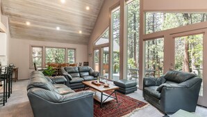 A spacious living room with leather sofas, a coffee table, and large windows offering a view of the forest. - Ponderosa Pine Escape  (Truckee)