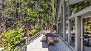 A deck with outdoor furniture, surrounded by tall trees and a glass building. - Ponderosa Pine Escape  (Truckee)