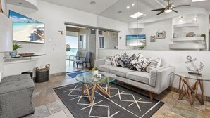 A modern living room with a glass coffee table, grey sofa, and a view of the ocean through a large window.