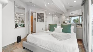 A modern bedroom with a large bed, a walk-in closet, a bathroom with a glass shower, and a view of palm trees.