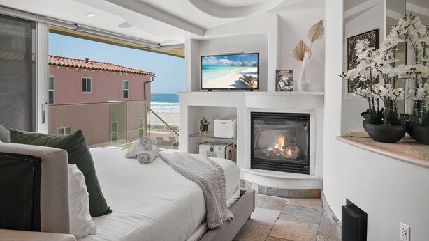 A modern bedroom with a large bed, a television, a fireplace, and a view of the beach.
