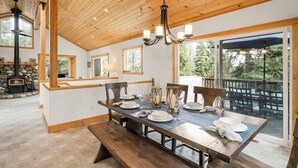 Un comedor con una mesa de madera dispuesta para cuatro, una chimenea de piedra y un gran ventanal corredizo a una terraza exterior.