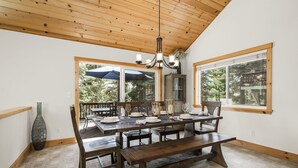 Un comedor con una mesa de madera dispuesta para seis comensales, una araña y grandes ventanales que ofrecen vistas al exterior.
