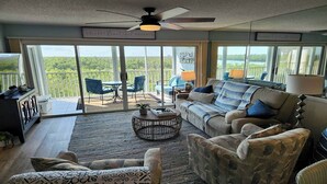 A spacious living room with a large sliding glass door leading to a balcony, a ceiling fan, and a view of greenery. - Beautiful 3/2 at Steven's Landing (Marco Island)