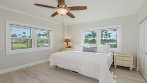 Une chambre à coucher comprenant un lit, des tables de chevet, un ventilateur de plafond et de grandes fenêtres donnant sur des palmiers.