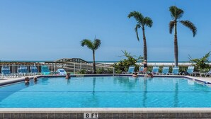 A swimming pool with clear blue water, surrounded by palm trees and lounge chairs, with a view of the ocean in the background.