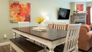 Een eetgedeelte met een houten tafel en stoelen, een vaas met gele bloemen, en een schilderij van rode bloemen aan de muur.