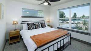 Une chambre à coucher comprenant un lit, des tables de chevet, un ventilateur de plafond, une fenêtre donnant sur un parking et une étagère fixée au mur.