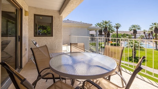 A glass-top table with chairs on a balcony with a view of a swimming pool and palm trees.