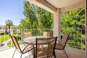A covered outdoor seating area with a round table and chairs, surrounded by a white railing, overlooking a landscaped garden with trees and a pathway.