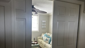 Ein Zimmer mit einem beigefarbenen Sofa, einem gemusterten Hocker, einem kleinen Tisch mit Lampe und einem Deckenventilator.