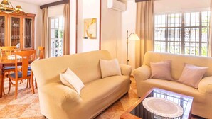 Ein Wohnzimmer mit zwei Sofas, einem Esstisch mit Stühlen, einem Couchtisch, einer Stehlampe und einem großen Fenster mit Vorhängen.