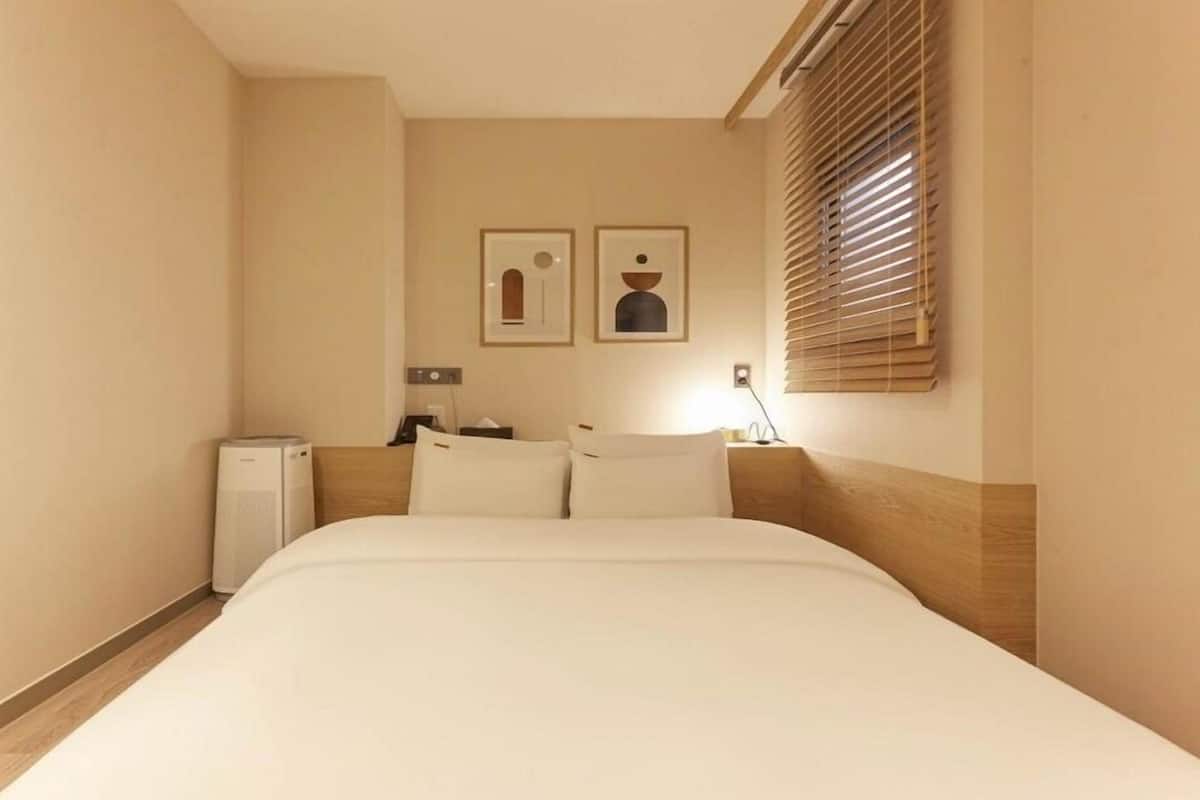 A hotel room with a bed, a bedside table, a lamp, a small heater, and two framed pictures on the wall.