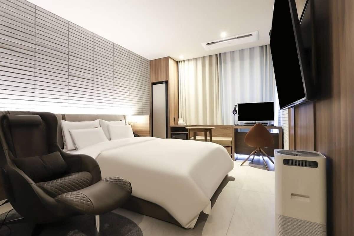 A modern hotel room with a large bed, a reclining chair, a desk, and a flat-screen TV.